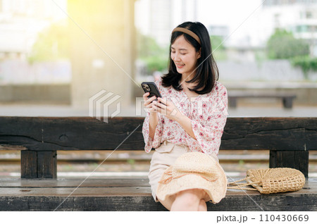 young asian woman traveler with weaving basket using a mobile phone and waiting for train in train station. Journey trip lifestyle, world travel explorer or Asia summer tourism concept. 110430669