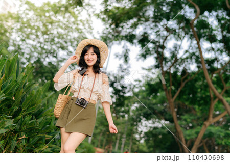 Portrait of asian young woman traveler with weaving hat and basket and a camera on green public park nature background. Journey trip lifestyle, world travel explorer or Asia summer tourism concept. 110430698