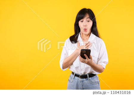 Surprised young asian woman using mobile phone with positive expression, smiles broadly, dressed in white shirt and standing isolated on yellow background. Happy adorable glad woman rejoices success. Surprised young asian woman using mobile phone with positive expression, smiles broadly, dressed in white shirt and standing isolated on yellow background. Happy adorable glad woman rejoices success. 110431016