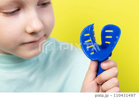 A seven year old girl looks at a blue dental impression tray in orthodontics. Concept for manufacturing braces and dentures for children, correction of malocclusion. Yellow background, dentistry 110431497