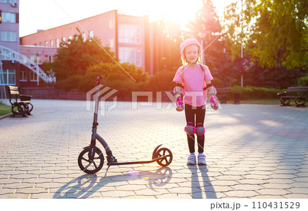 A little caucasian girl in a pink protective gear laughs and grimaces near a scooter against the backdrop of a summer sunset. 110431529