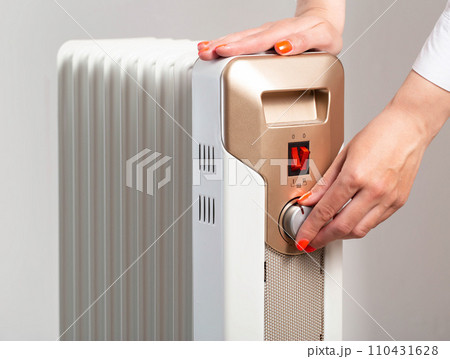 A woman s hand with a beautiful manicure regulates the room heating temperature on an electric heater. The concept of turning on the heating in the autumn-winter period. Cold in apartments, close-up 110431628