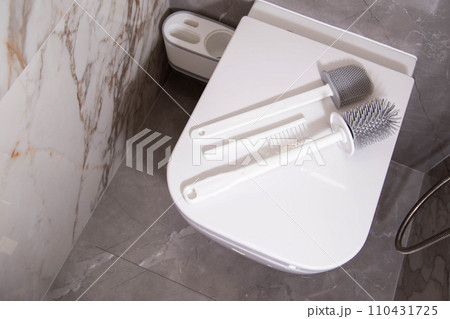 Silicone brushes for cleaning the toilet lie on the lid of the wall-hung toilet. The concept of care and cleaning the toilet from dirt and bacteria. Gray stoneware tiles on the walls. Copy space for Silicone brushes for cleaning the toilet lie on the lid of the wall-hung toilet. The concept of care and cleaning the toilet from dirt and bacteria. Gray stoneware tiles on the walls. Copy space for 110431725