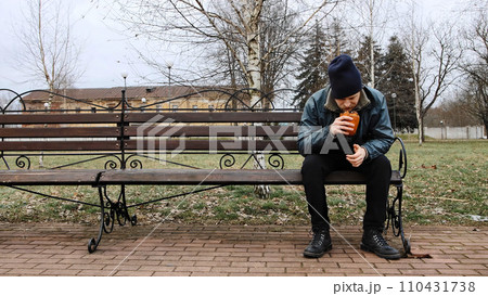 Hungry bearded homeless man sits on a bench and eats a bun in a city park. Below poverty line. Male tramp in dirty clothes with cap and hat. Beggar. Illegal immigrant. Drunkenness, alcoholism Hungry bearded homeless man sits on a bench and eats a bun in a city park. Below poverty line. Male tramp in dirty clothes with cap and hat. Beggar. Illegal immigrant. Drunkenness, alcoholism 110431738
