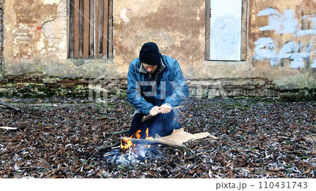 Homeless man sits on ground and pushes pieces of cardboard into fire to keep warm from cold. Tramp in dirty clothes and hat cap. Below poverty line. Illegal immigrant. Male refugee. Fugitive beggar 110431743