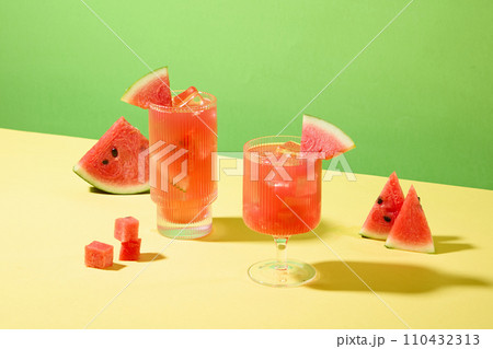 Close-up of two glasses of watermelon juice with ice and fresh watermelon slices on a green-yellow background. Supplement vitamins from fresh fruits. 110432313