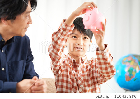 貯金箱にお金を入れる小学生とその父親 110433886