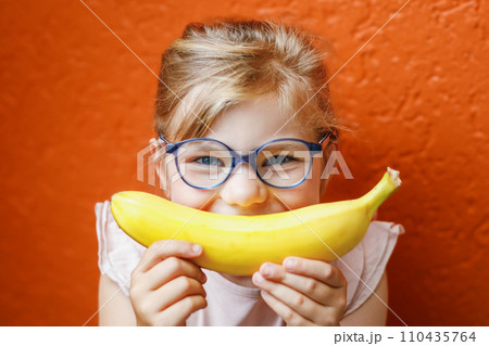 Happy little child girl with yellow banana like smile on orange background. Preschool girl with glasses smiling. Healthy fruits for children Happy little child girl with yellow banana like smile on orange background. Preschool girl with glasses smiling. Healthy fruits for children 110435764