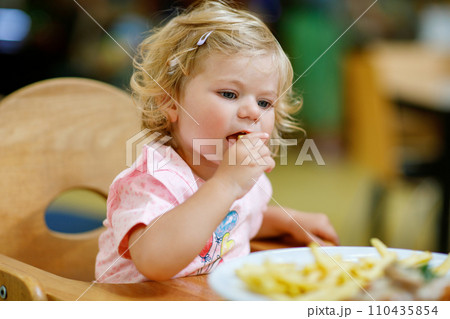 Adorable toddler girl eating healthy vegetables and unhealthy french fries potatoes. Cute happy baby child taking food from dish at daycare or nursery canteen. 110435854