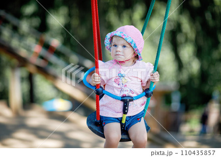 Cute adorable toddler girl swinging on outdoor playground. Happy smiling baby child sitting in chain swing. Active baby on sunny summer day outside. Family leisure and activity outdoors. Cute adorable toddler girl swinging on outdoor playground. Happy smiling baby child sitting in chain swing. Active baby on sunny summer day outside. Family leisure and activity outdoors. 110435857