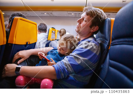 Young father holding his baby toddler daughter during flight on airplane going on vacations. Tired man and cute girl during night flight. child playing with smartphone. Family sitting in plane Young father holding his baby toddler daughter during flight on airplane going on vacations. Tired man and cute girl during night flight. child playing with smartphone. Family sitting in plane 110435863