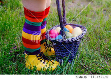 Close-up of legs of toddler girl with colorful stockings and shoes and basket with colored eggs. Child having fun with traditional Easter eggs hunt, outdoors. Celebration of christian holiday. Close-up of legs of toddler girl with colorful stockings and shoes and basket with colored eggs. Child having fun with traditional Easter eggs hunt, outdoors. Celebration of christian holiday. 110435920