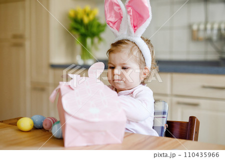 Cute little toddler girl wearing Easter bunny ears playing with colored pastel eggs. Happy baby child unpacking gifts. Adorable healthy smiling kid in pink clothes enjoying family holiday Cute little toddler girl wearing Easter bunny ears playing with colored pastel eggs. Happy baby child unpacking gifts. Adorable healthy smiling kid in pink clothes enjoying family holiday 110435966