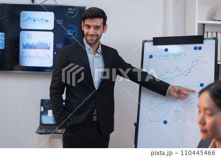 A South Asian man in a business suit points to growth charts during a dynamic corporate strategy session, confidently leading the discussion. 110445466