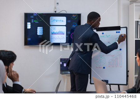 An African man in a navy suit actively engages with charts on a whiteboard, leading a strategic business meeting with confidence and expertise. 110445468