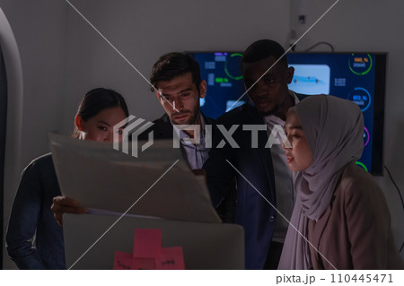 A multicultural team, including an Asian woman, a South Asian man, an African man, and a woman in a hijab, closely review a document, showcasing collaboration and focus in a business environment. 110445471