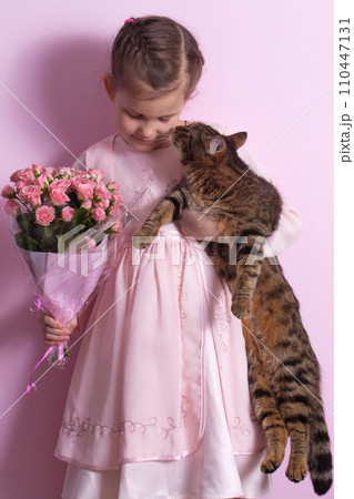 A cute little girl and a cat in a pink dress holds a bouquet of pink roses A cute little girl and a cat in a pink dress holds a bouquet of pink roses 110447131