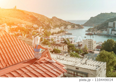 Woman sits on rooftop, enjoys town view and sea mountains. Peaceful rooftop relaxation. Below her, there is a town with several boats visible in the water. Rooftop vantage point. Woman sits on rooftop, enjoys town view and sea mountains. Peaceful rooftop relaxation. Below her, there is a town with several boats visible in the water. Rooftop vantage point. 110449014
