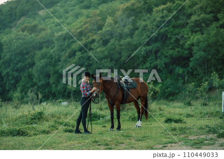 Happy blonde with horse in forest. Woman and a horse walking through the field during the day. Dressed in a plaid shirt and black leggings. 110449033
