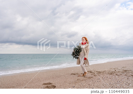 Redhead woman Christmas tree sea. Christmas portrait of a happy redhead woman walking along the beach and holding a Christmas tree in her hands. Dressed in a light coat, white suit and red mittens. 110449035