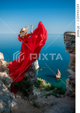 Red dress sea woman. Happy woman with flowing hair in a long flowing red dress stands on a rock near the sea. Travel concept, photo session at sea 110449184