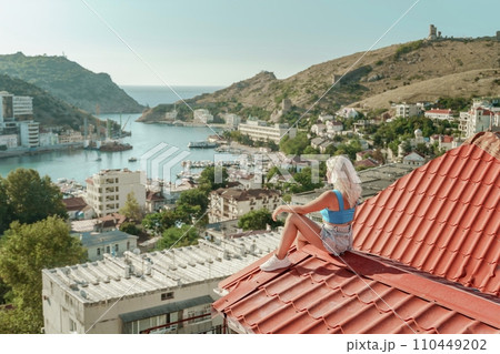Woman sits on rooftop, enjoys town view and sea mountains. Peaceful rooftop relaxation. Below her, there is a town with several boats visible in the water. Rooftop vantage point. Woman sits on rooftop, enjoys town view and sea mountains. Peaceful rooftop relaxation. Below her, there is a town with several boats visible in the water. Rooftop vantage point. 110449202
