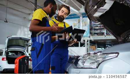Repair shop employees work together on fixing car, using tablet and laptop. Teamworking colleagues collaborating on servicing broken vehicle, checking for defective components using electronic devices 110456587