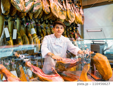 Male seller cutting slices from whole leg of jamon in meat store 110456627