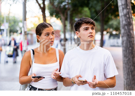 Young guy and girl using mobile phones while walking along summer city street Young guy and girl using mobile phones while walking along summer city street 110457018