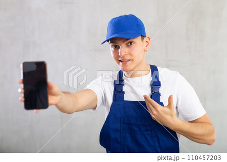 Positive man in blue protective overalls showing modern smart phone with blank screen Positive man in blue protective overalls showing modern smart phone with blank screen 110457203