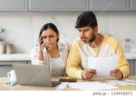 Overwhelmed couple sitting together at kitchen, using laptop, holding papers Overwhelmed couple sitting together at kitchen, using laptop, holding papers 110458871