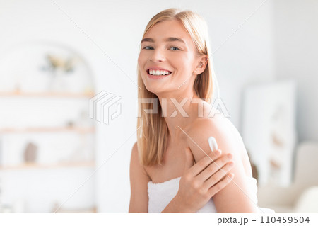 Young blonde woman applying moisturizer cream to shoulders skin indoors 110459504
