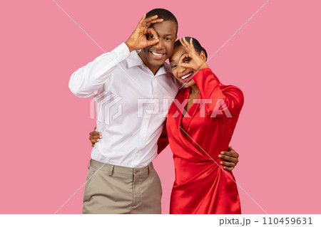 Black couple making fun hand gestures, woman in red, man in white, pink background 110459631