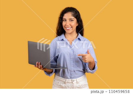 Friendly young woman with curly hair holds a laptop and points to it with a smile 110459658