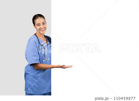 Inviting european nurse in blue scrubs presenting with an open hand gesture beside a blank space 110459877