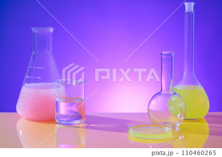 Erlenmeyer flask, boiling flask, beaker and transparent petri dish displayed with colored liquid on a pink-purple gradient background. Blank space with front view. 110460265