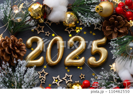 The golden figures 2025 made of candles on a black stone slate background are decorated with a festive decor of stars, sequins, fir branches, balls and garlands. Greeting card, happy New Year. 110462550