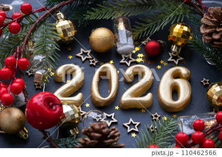 The golden figures 2026 made of candles on a black stone slate background are decorated with a festive decor of stars, sequins, fir branches, balls and garlands. Greeting card, happy New Year. 110462566