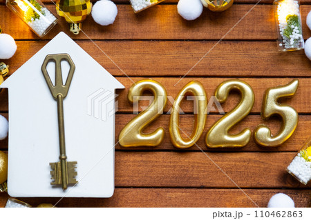 House key with tiny figure of home mock up on festive brown wooden background, lights of garlands. New Year 2025 wooden letters, greeting card. Purchase, construction, relocation, mortgage, insurance 110462863