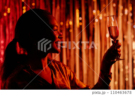 Side view of cheerful girl in red dim light having fun at dance club party while looking at glass of champagne in raised hand Side view of cheerful girl in red dim light having fun at dance club party while looking at glass of champagne in raised hand 110463678