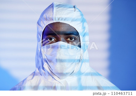 Close up shot of African American scientist looking at camera dressed in white hazmat suit and mask while standing in blue light and blinds shadow 110463711