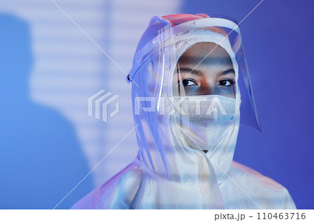 Medium close up shot female laboratory technician dressed in hazmat suit, face shield and mask looking at camera while standing at blue background with blinds shadow Medium close up shot female laboratory technician dressed in hazmat suit, face shield and mask looking at camera while standing at blue background with blinds shadow 110463716