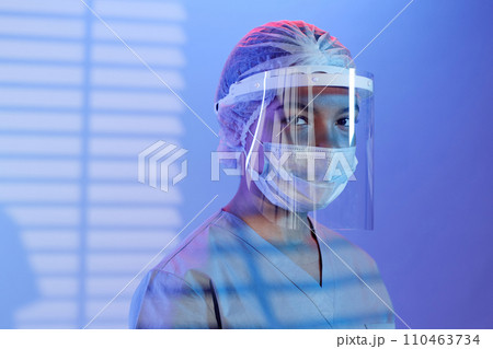 Medium close up shot confident woman surgeon wearing medical protective wear looking at camera while standing in blue light and shadow from window blinds 110463734