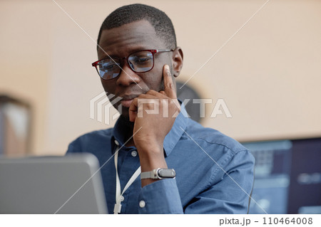 Medium close up shot of thoughtful African American male IT company employee wearing glasses working on laptop at table Medium close up shot of thoughtful African American male IT company employee wearing glasses working on laptop at table 110464008