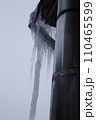 A huge ice icicle hangs on the edge of the roof of a multi-story building. An icicle hangs near a metal pipe for ventilation. 110465599