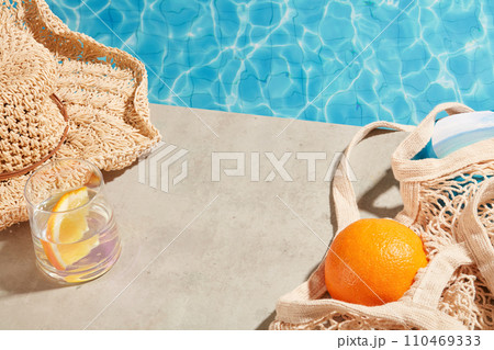 Summer vacation by the water with a mesh bag, beach hat and a cup filled with water and slice of orange inside. Beauty product can be displayed in the vacant space 110469333