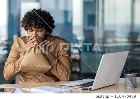 Panic attack in worker inside office at workplace, businessman breathing hard using paper bag, working sitting with laptop. 110470414