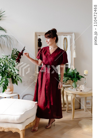 Adult woman, dressed in red dress, stands in classic light living room 110472468