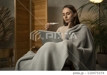 A sick woman, wrapped in a blanket, conducts an online appointment with a virtual doctor using a laptop 110473374