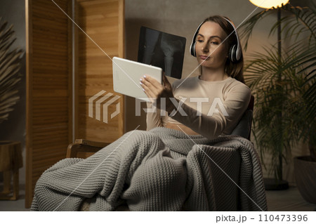 Beautiful woman is making a video call with her doctor in headphones while sitting on an armchair. The woman shows the doctor an X-ray. 110473396